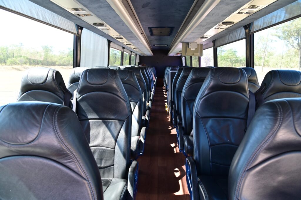 Rows of empty, cushioned leather seats are lined on both sides of the aisle inside a bus. Sunlight filters through large windows, revealing a wooded area outside.