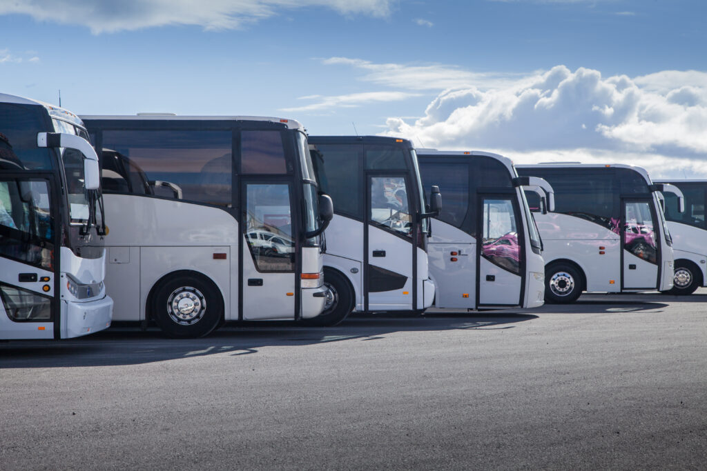 Buses parked in a neat row, facing right, under a bright blue sky with scattered clouds. The scene suggests a transportation hub or rest area.