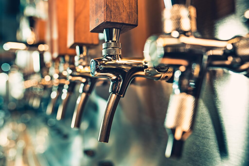 Metal beer taps with wooden handles are lined up closely, ready to pour beverages in a bar setting, reflecting ambient light on the metallic surface.