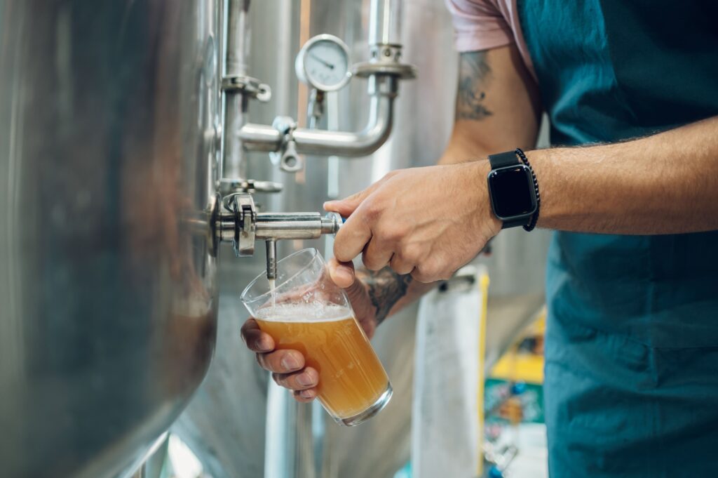 A person pours beer from a stainless steel tank into a glass, wearing an apron and smartwatch, in an industrial setting with pipes and gauges.