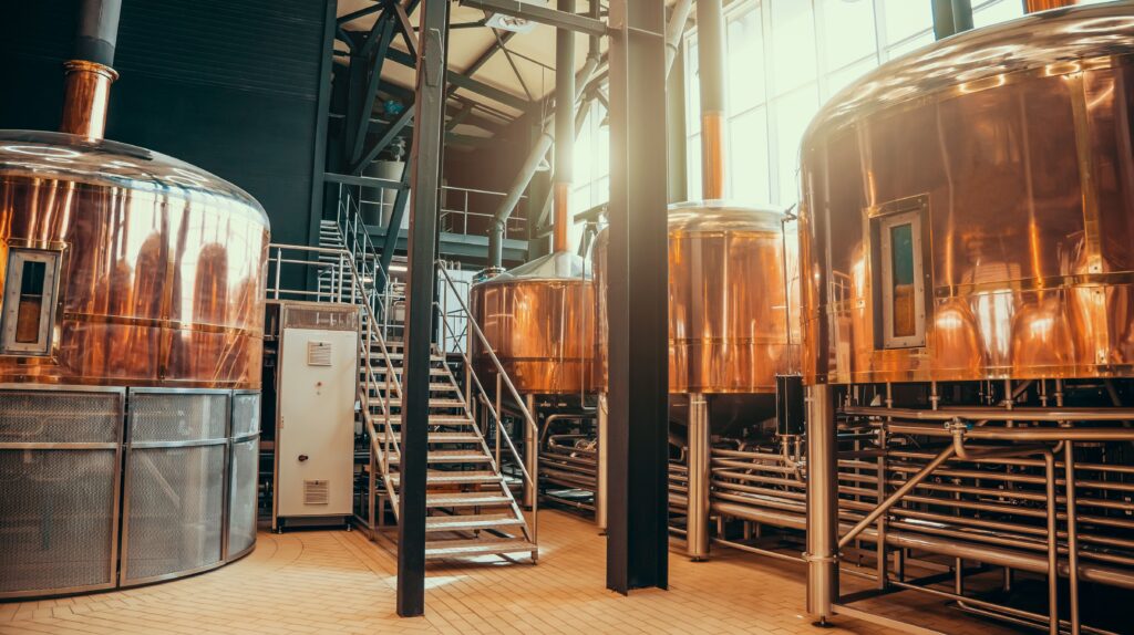 Copper brewing tanks stand in a brewery, surrounded by pipes and scaffolding. Metal stairs lead to an elevated platform, with sunlight streaming through large windows, illuminating the industrial scene.