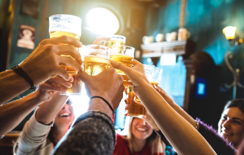 Hands holding glasses of beer are raised in a toast, surrounded by smiling faces in a warmly lit bar with teal walls and wooden decor.