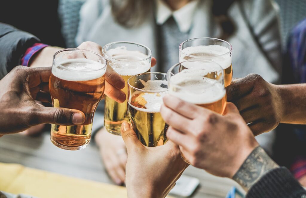 Hands clink beer glasses in a celebratory toast, with foamy golden liquid inside. The setting appears social, with people gathered closely, suggesting a lively gathering or celebration.
