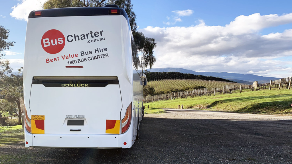 A white bus labeled "Bus Charter Best Value Bus Hire 1800 BUS CHARTER BONLUCK" is parked on a gravel path, surrounded by lush vineyards and hills under a partly cloudy sky.