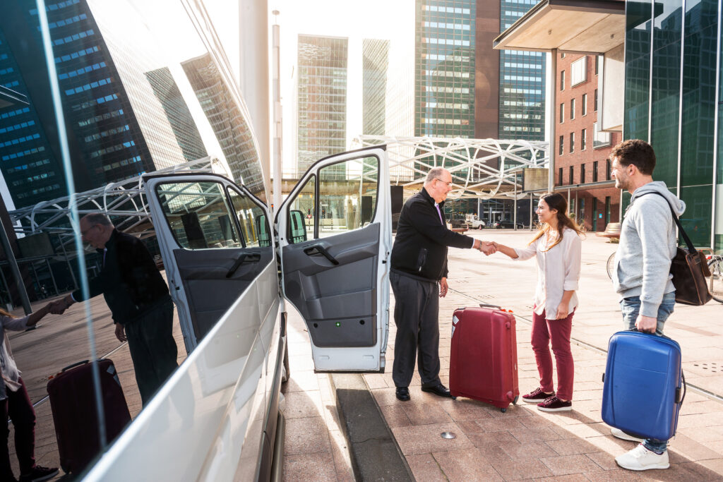 A man shakes hands with a woman while standing next to a white van. Both carry suitcases, with urban buildings in the background, suggesting a travel or transportation context.