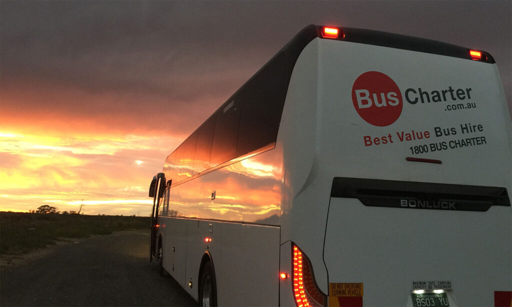 A white bus labeled "Bus Charter .com.au" and "Best Value Bus Hire 1800 BUS CHARTER" is parked on a road. It faces a vivid sunset, casting warm light on the scene.