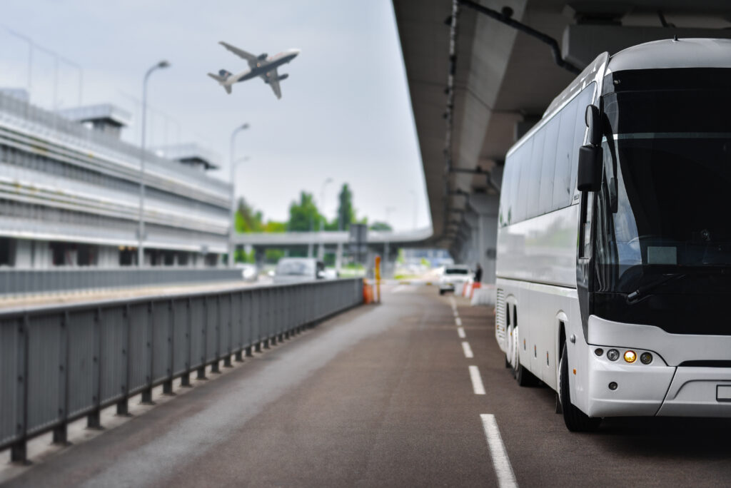 A white bus parked under an overpass at an airport terminal, with an airplane flying overhead and adjacent buildings visible in the background.