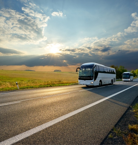 two white buses travel on a country road at sunset, surrounded by vast fields and a dramatic, cloudy sky.