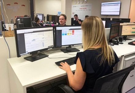 a woman works at a desk with three computer monitors displaying software applications. in the office setting, colleagues sit and work at their desks, with additional monitors mounted on the walls.